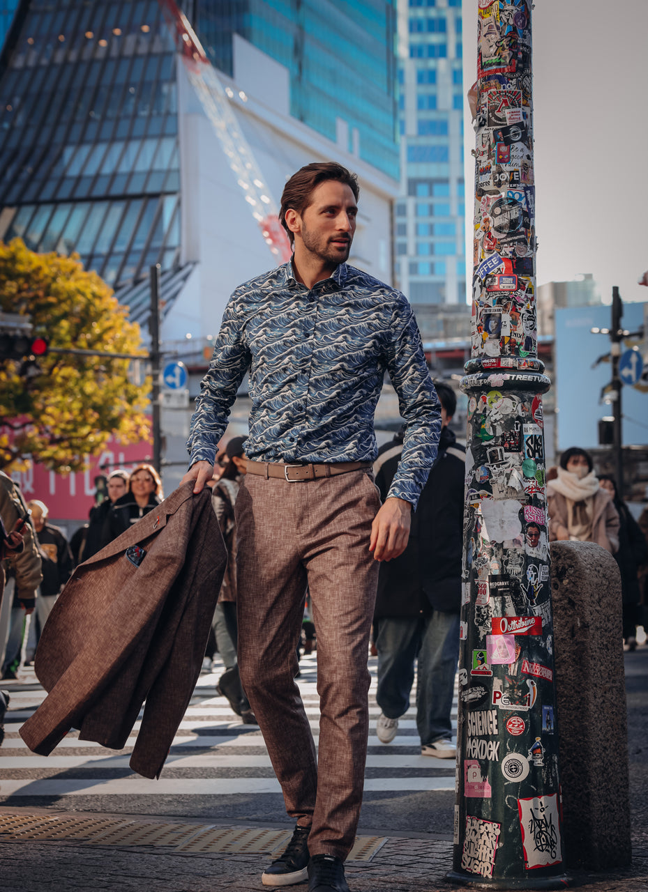 A man walking on a city street wearing a blue and gray patterned button-down shirt, brown trousers, and holding a brown blazer. He is walking next to a pole covered in stickers.