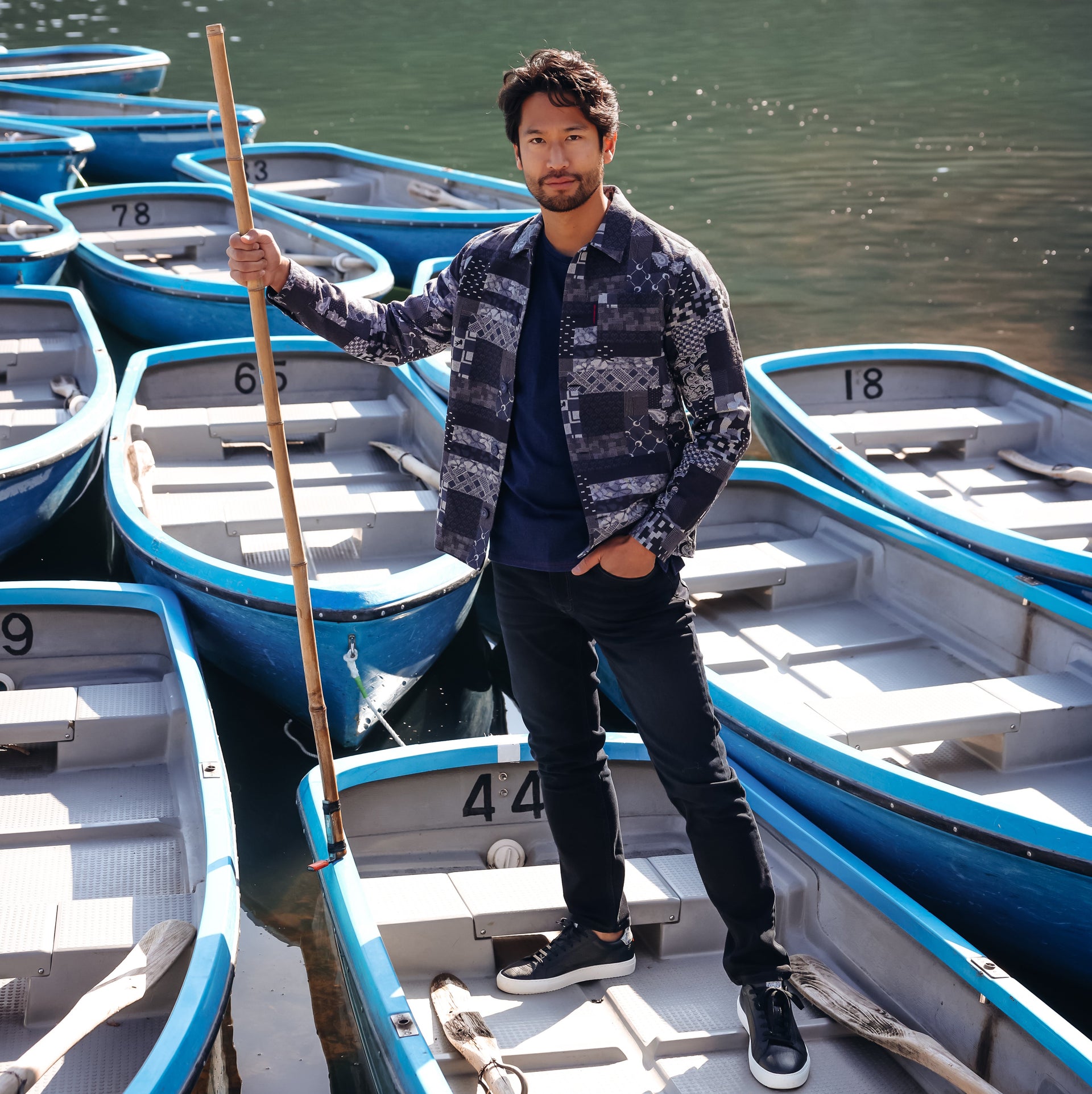 A man stands in a blue and white rowboat on a dock, holding a wooden paddle, with multiple similar boats surrounding him on a calm lake.