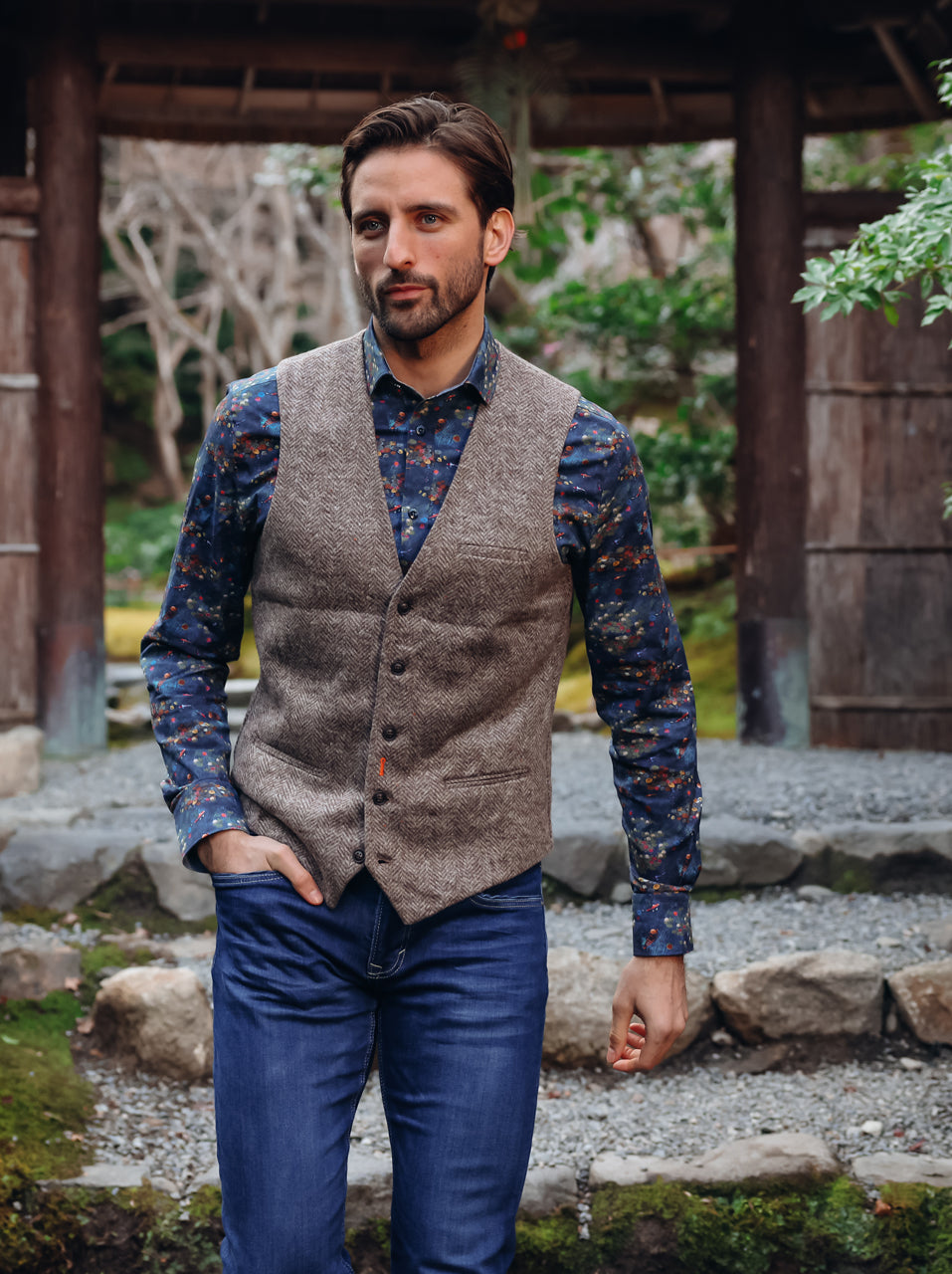 A Fish Named Fred men's Brushed Herringbone waistcoat in Sand, worn by a model at a traditional garden gate