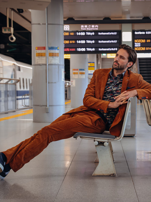 A Fish Named Fred men's Rib trousers in Cognac, worn by a model seated on a station platform bench