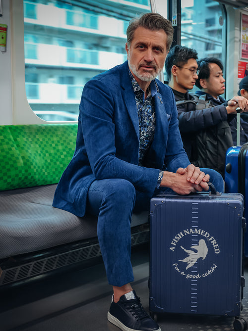 A Fish Named Fred men's Rib trousers in Night Sky Blue, worn by a model seated on a train with luggage