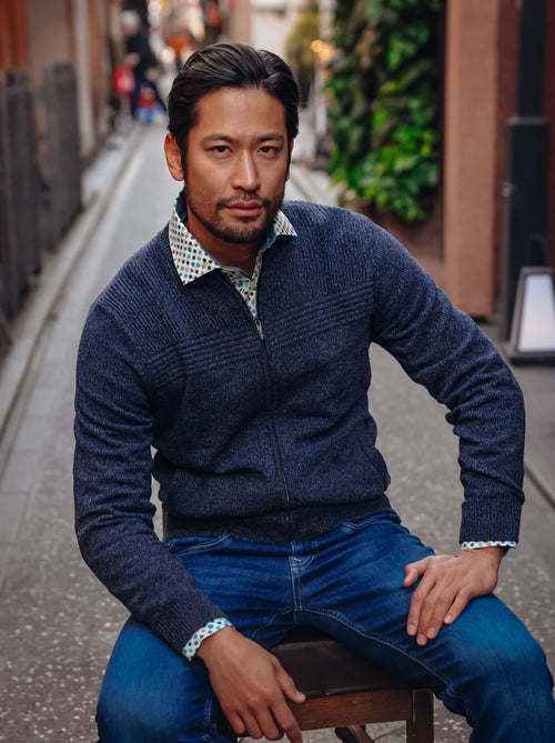 A Fish Named Fred men's Melange cardigan in Navy, worn by a model seated on a stool in an alley