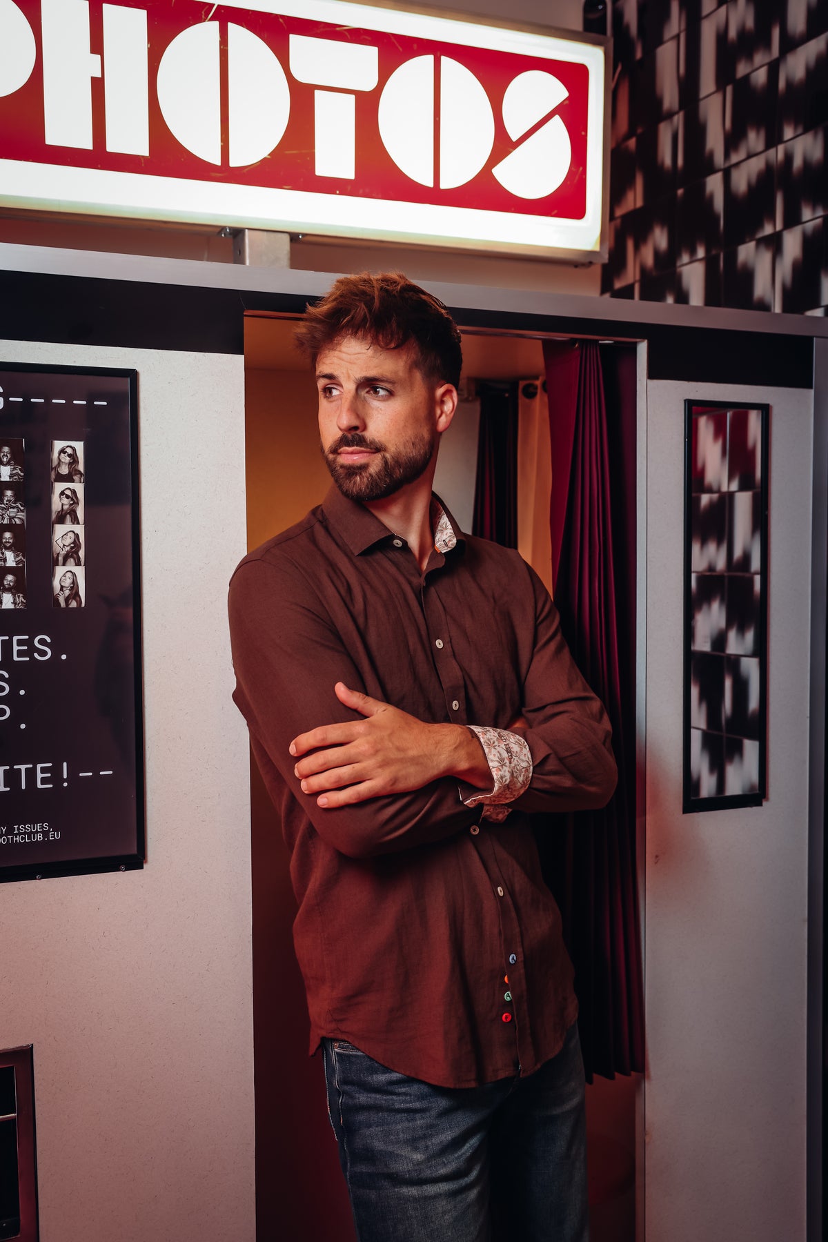 A man with a beard stands with arms crossed in a doorway, wearing a brown button-up shirt and blue jeans. A red and white sign reading 'PHOTOS' is visible above him, with framed wall art and curtains in the background.