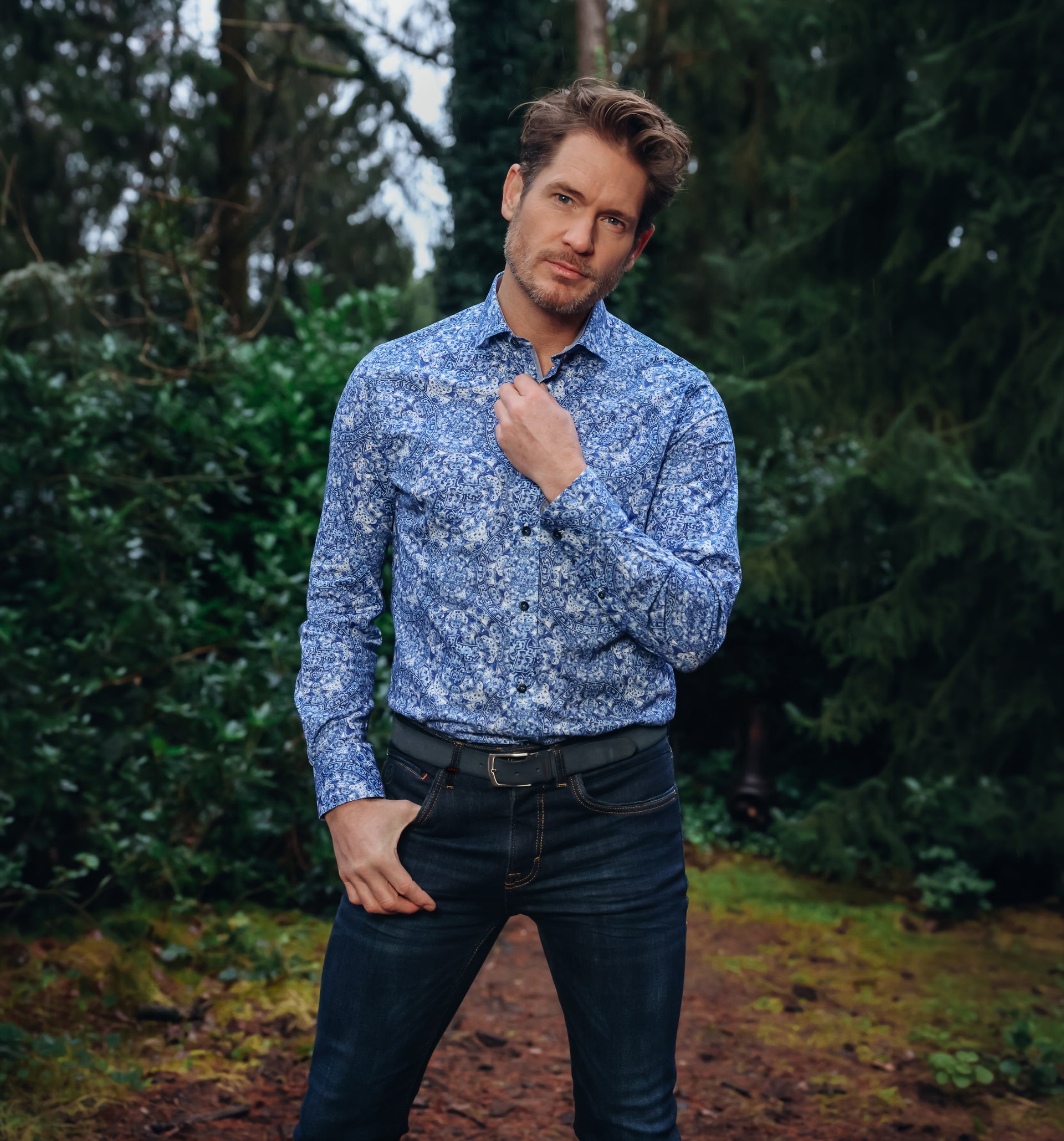 Man wearing a blue floral shirt and dark jeans standing in a garden.
