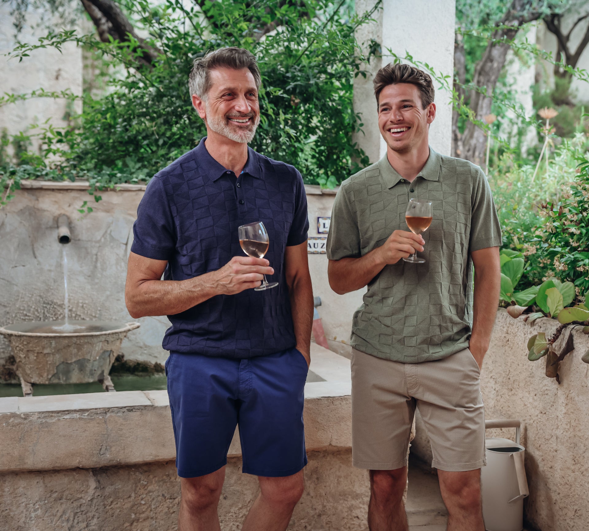 Two men standing outdoors, holding glasses of wine, with a garden and fountain in the background.