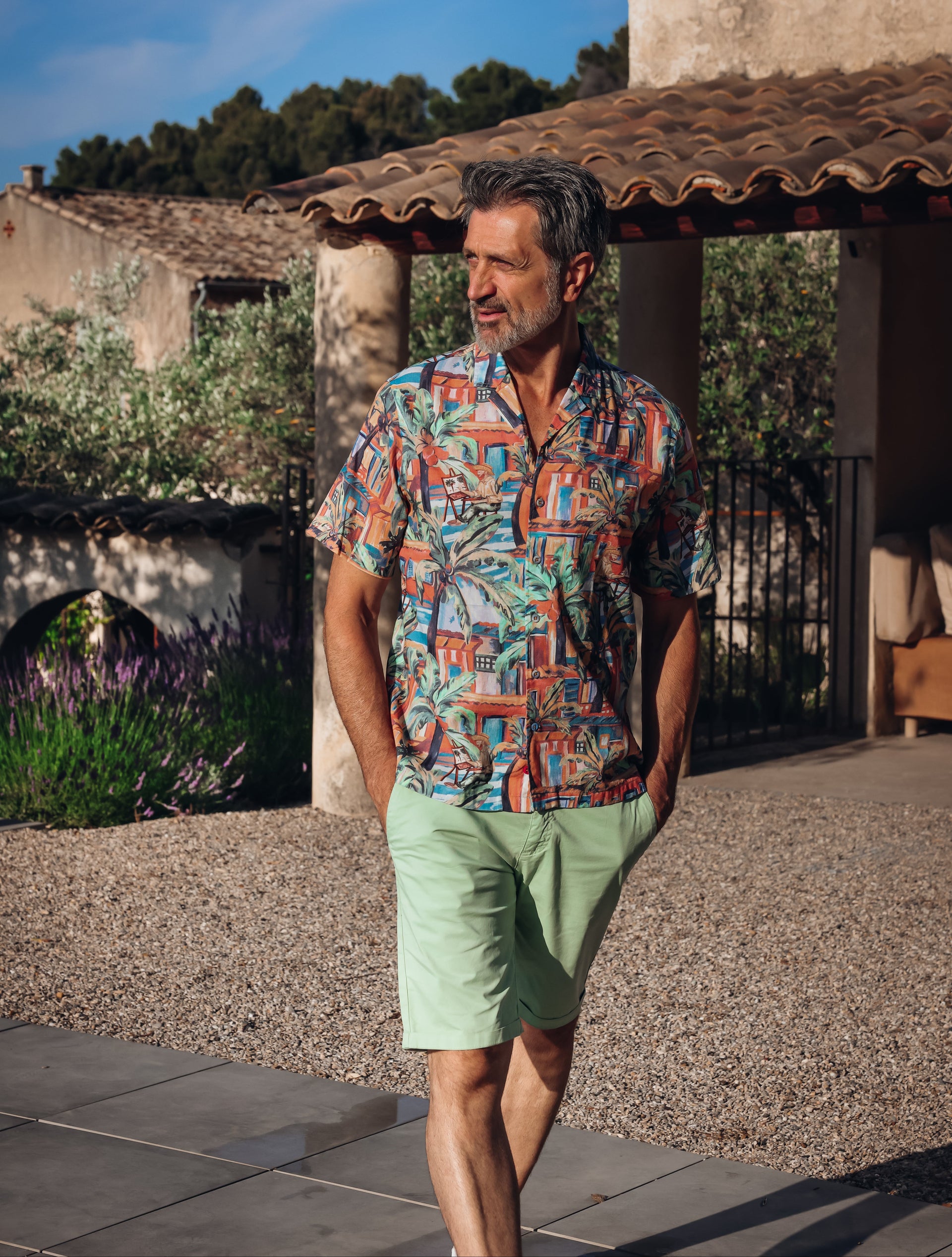 A man wearing a short-sleeve button-up shirt with a colorful tropical and geometric print, paired with light green shorts, walking outdoors in front of a rustic stone building under a blue sky.
