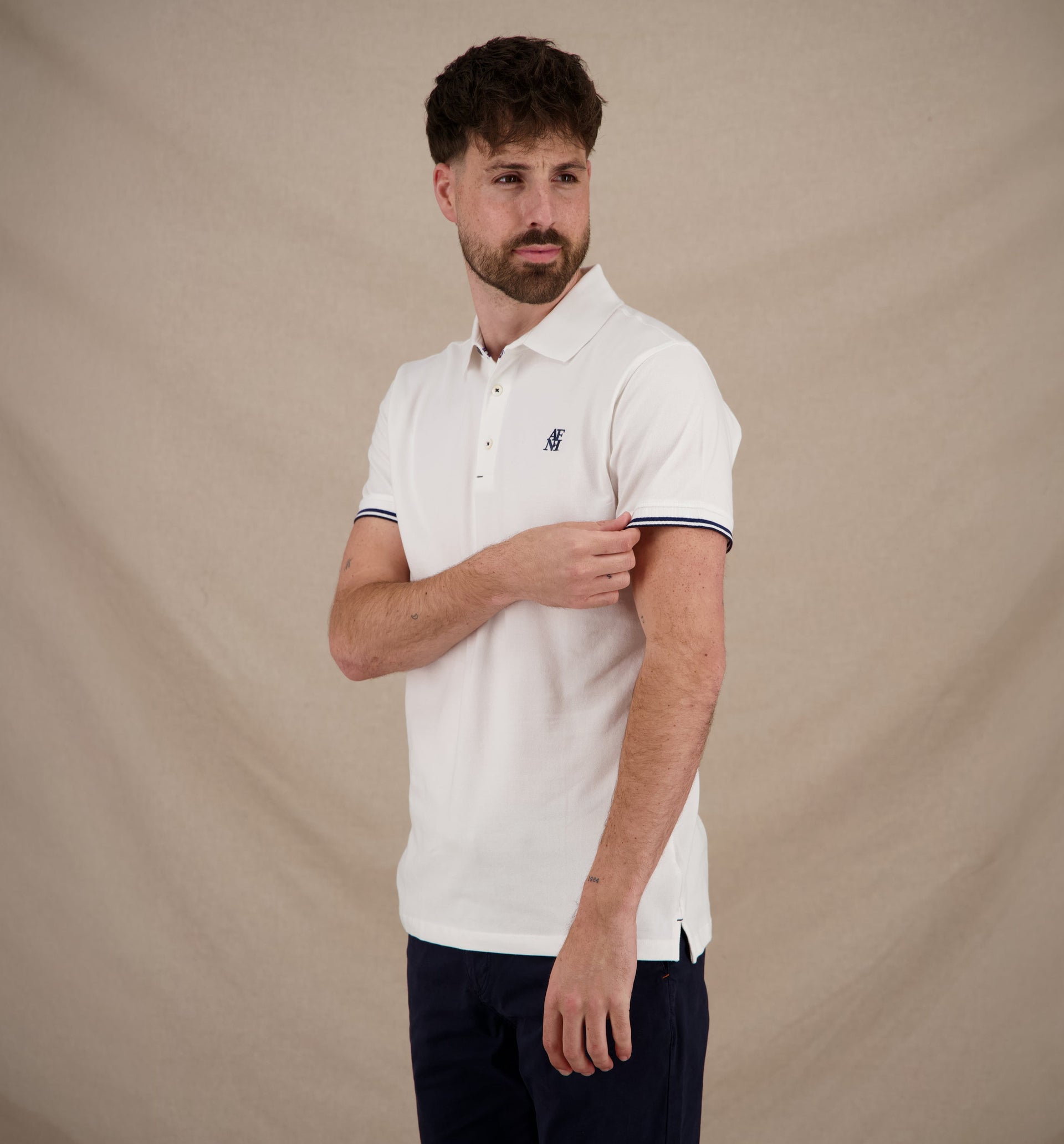 A man wearing a white short-sleeve polo shirt with navy and white striped trim on the sleeve and a small embroidered logo on the chest, paired with dark navy trousers, standing against a beige fabric backdrop.
