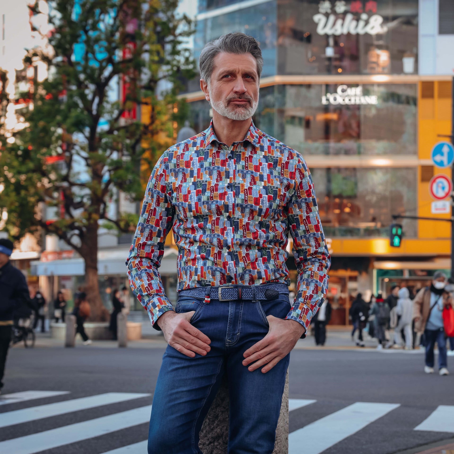 A man wearing a long-sleeve button-down shirt with a colorful geometric block pattern in red, blue, yellow, and white, paired with dark blue jeans, standing on a city street crosswalk.