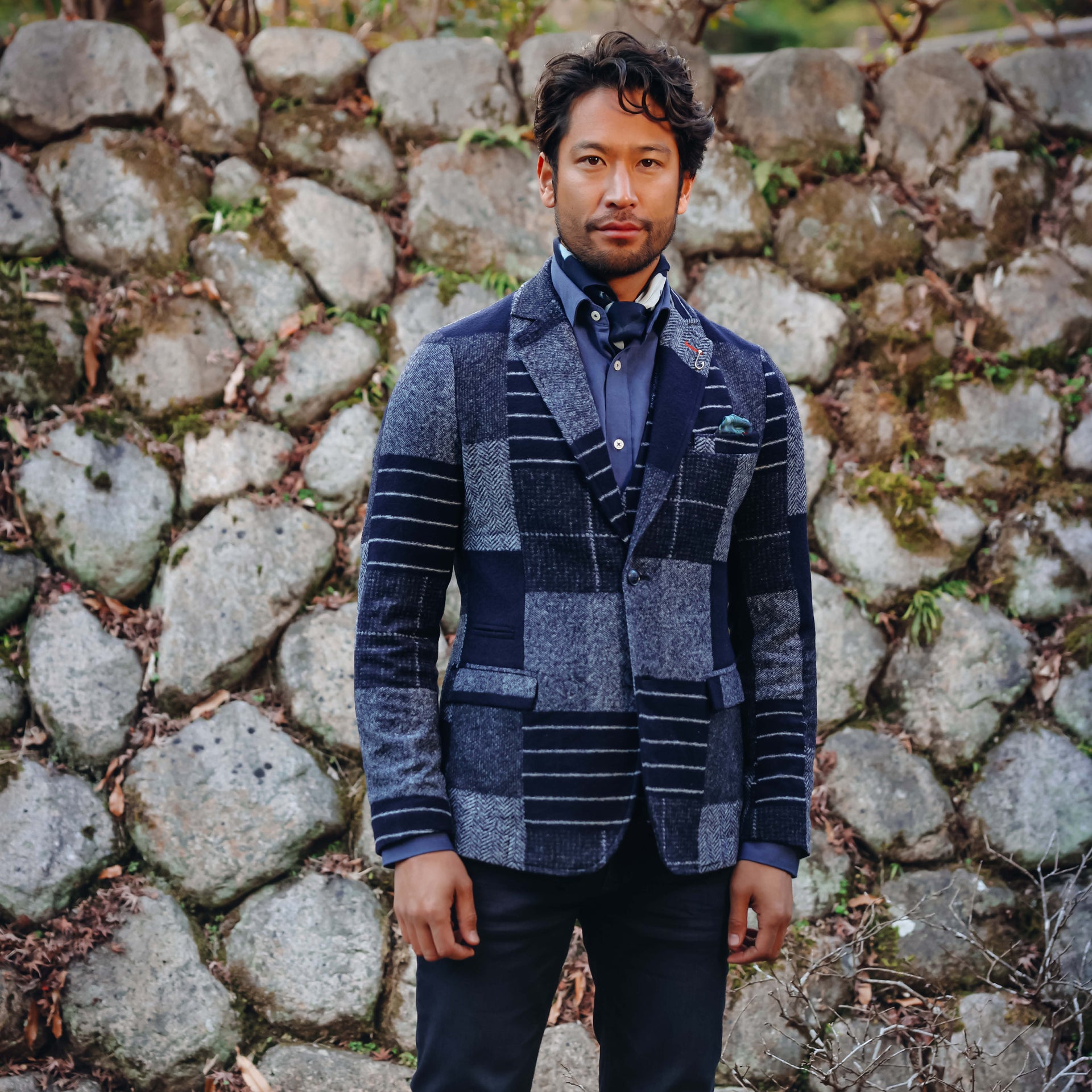A man wearing a navy and gray patchwork blazer with a patterned pocket square, standing in front of a rustic stone wall.