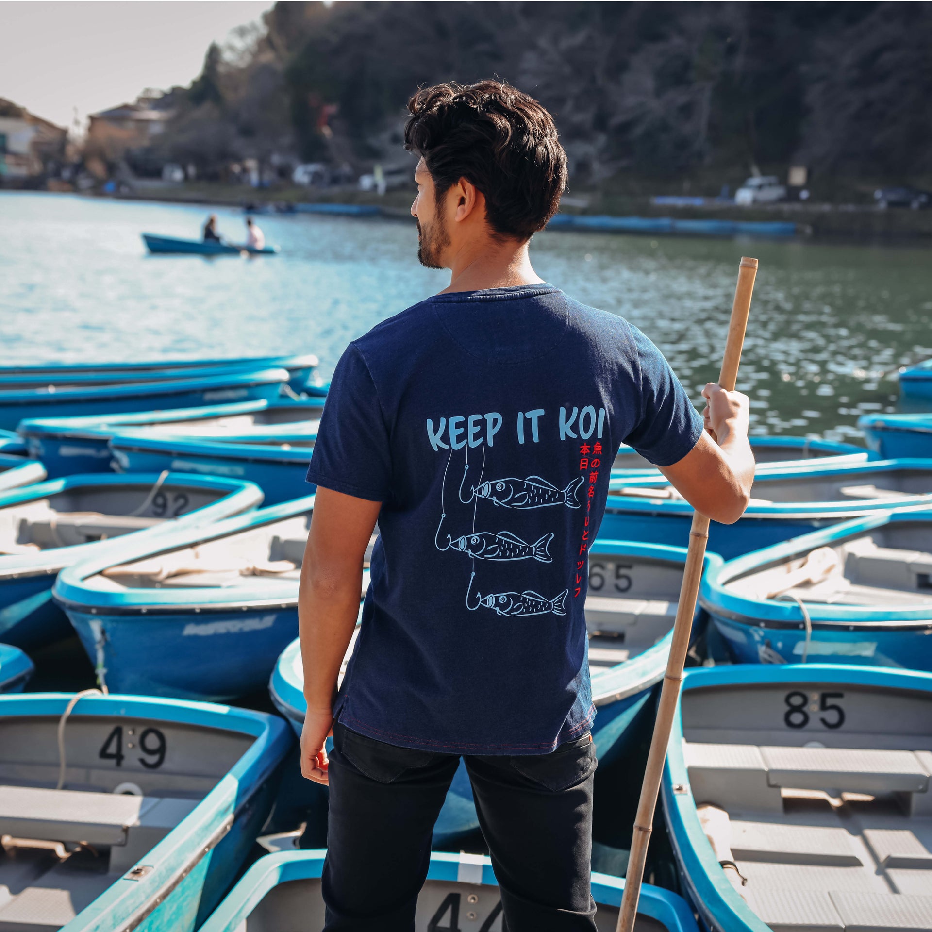 A man stands on a dock holding a wooden paddle, viewed from behind, wearing a navy blue t-shirt with a white graphic of three fishing lines and fish and the text 'KEEP IT KOI'. The background features numerous blue and white rowboats on the water.