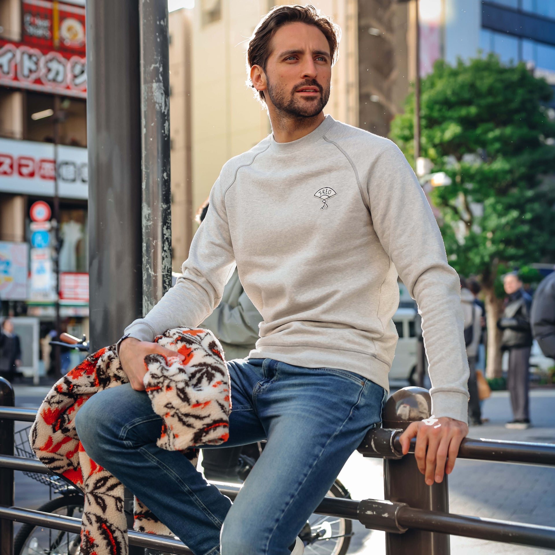 A man wearing a light beige crewneck sweatshirt with a small embroidered graphic on the chest, paired with blue jeans, sitting on a metal railing in an outdoor urban setting.