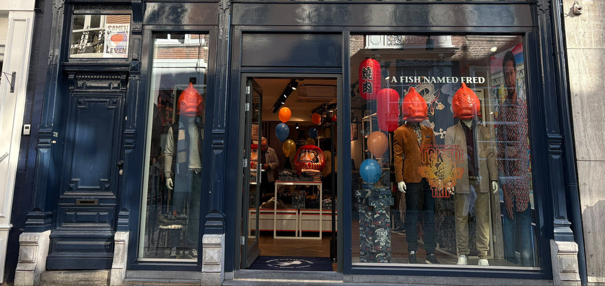 A storefront facade of a dark navy blue shop with large windows displaying mannequins wearing colorful garments. The building has cream-colored upper windows and stone detailing, with a street setting in front.