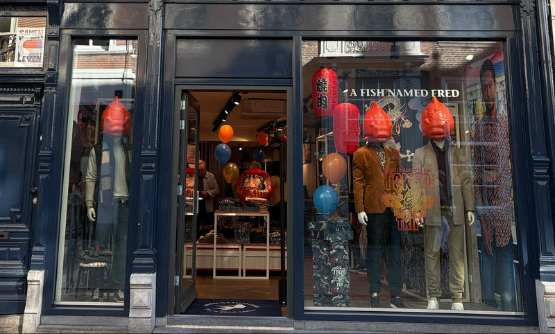A storefront facade of a dark navy blue shop with large windows displaying mannequins wearing colorful garments. The building has cream-colored upper windows and stone detailing, with a street setting in front.