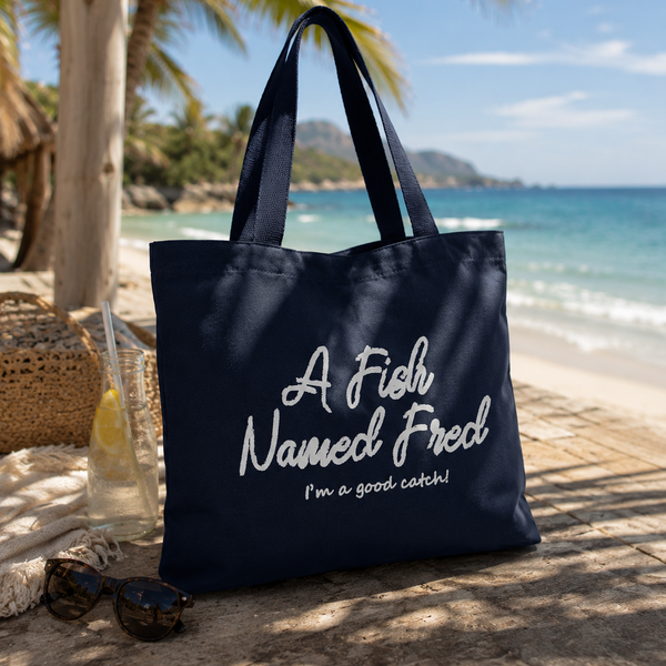 A navy blue canvas tote bag with white text reading 'A Fish Named Fred I'm a good catch!' is placed on a sandy beach with palm trees, ocean, sunglasses, and a drink in the background.